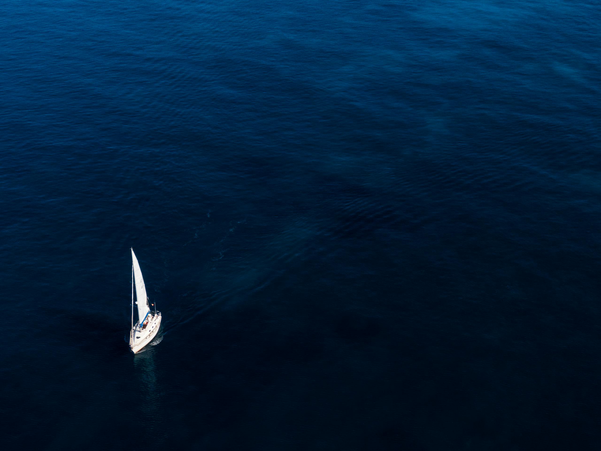 A lone sailboat glides on the vast, blue ocean under clear skies.